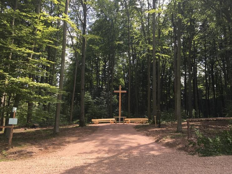 Holz-Kreuz auf einem gepflasterten Weg umgeben von B&auml;umen in einem lichtdurchfluteten Wald. Sitzb&auml;nke sind ebenfalls vorhanden.