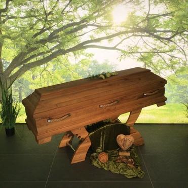 Holz-Sarg auf einem Tisch, umgeben von grünem Stoff und künstlichen Pflanzen, vor einem Hintergrund mit Bäumen und Licht.