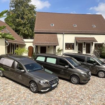 Drei Wagen, darunter ein Bestattungswagen, parken gepflastertem Hof vor einem Wohngebäude mit brauner Dachdeckung.