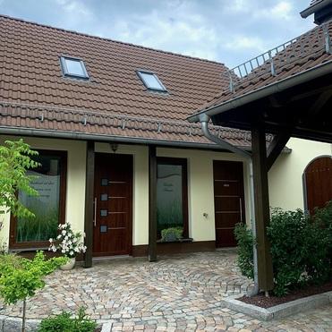 Eingangsbereich eines Hauses mit zwei Holztüren und einer gepflasterten Auffahrt umgeben von Pflanzen und Bäumen.