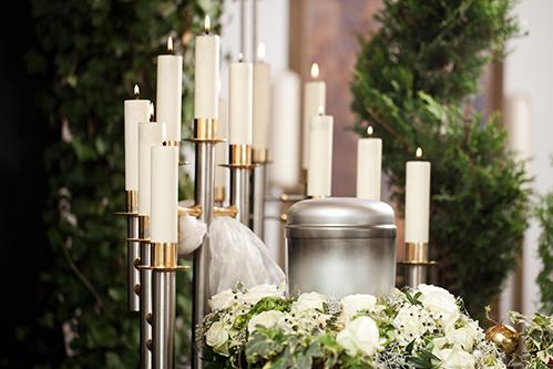 Grabschmuck mit wei&szlig;en Rosen und Kerzen um eine silberne Urne auf einem Tisch.