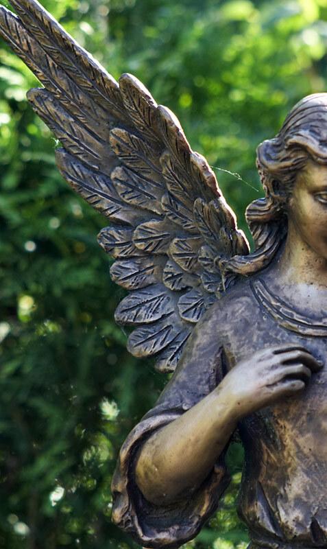 Bronzefarbene Engelstatue mit detaillierten Fl&uuml;geln, umgeben von gr&uuml;nem Hintergrund.