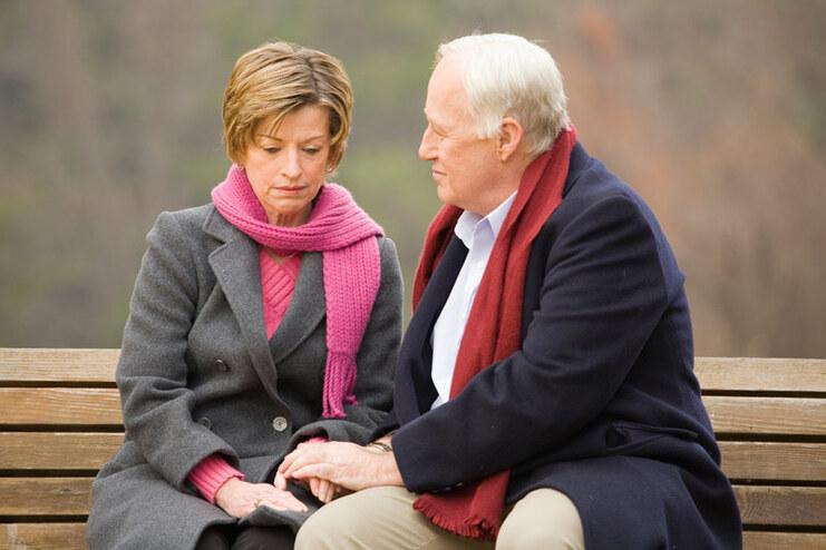 Ein Paar sitzt auf einer Bank, die Frau tr&auml;gt einen grauen Mantel und Schal, der Mann h&auml;lt ihre Hand und schaut sie an.