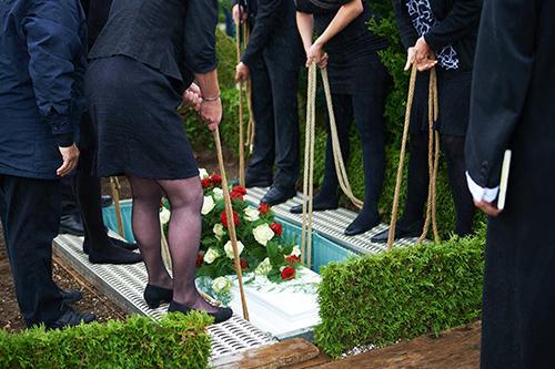 Menschen in schwarzer Kleidung senken ein Blumengesteck &uuml;ber einem offenen Grab. Erde und Strick sind sichtbar.