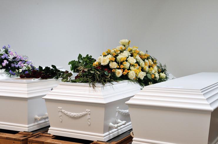 Drei weiße Särge mit verschiedenen Blumenarrangements, darunter gelbe und rote Rosen, auf Holzkisten.