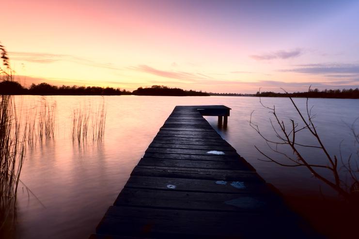 Holzsteg führt über ruhiges Wasser, umgeben von Gräsern und einem farbenprächtigen Sonnenuntergang.