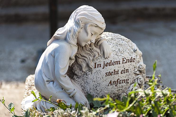 Skulptur eines mädchenhaften Engels, der auf einem Grabstein mit der Inschrift steht: "In jedem Ende steht ein Anfang."