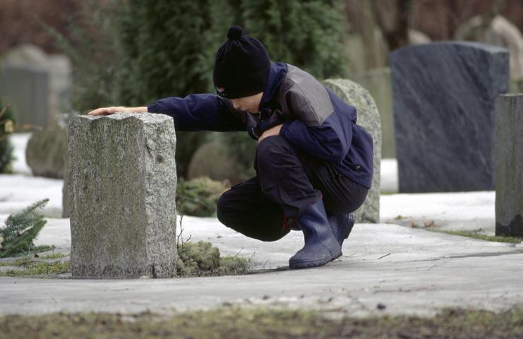 Ein Kind kniet neben einem Grabstein auf einem Friedhof und berührt nachdenklich die Oberfläche des Steins.