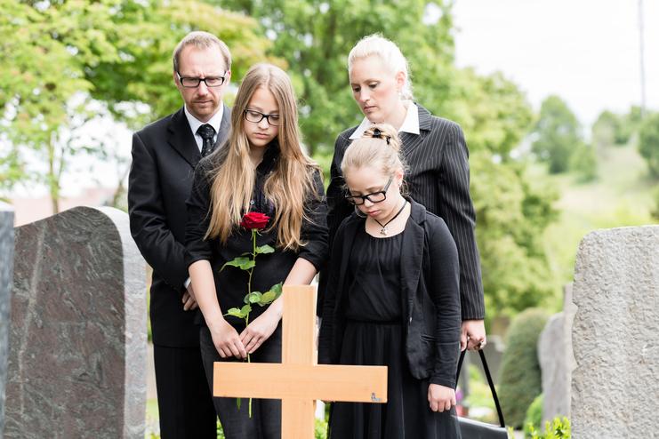 Familie in Trauerkleidung steht an einem Grab, während eine Jugendliche eine Rose hält und den Kopf senkt.